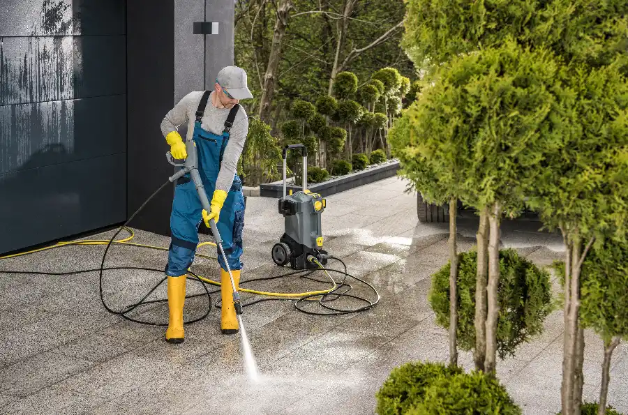 A person in blue overalls and yellow rubber boots and gloves uses a pressure washer to clean a concrete paver driveway, with a portable pressure washer unit and trimmed shrubs in the background.